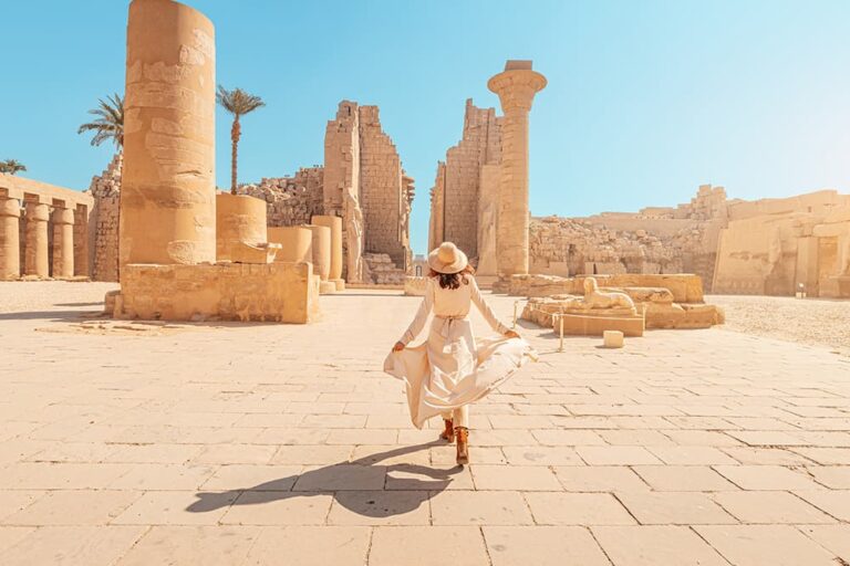 エジプトを代表する観光地であるルクソールのカルナック神殿にて、青空の下、巨大な石柱や遺跡群の間を歩く女性観光客の後ろ姿