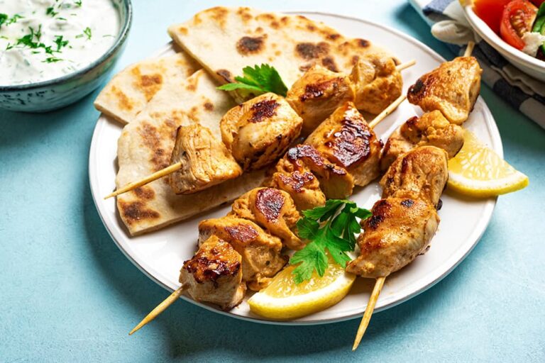 ヨーロッパで人気の食べ物であるギリシャの代表的な肉料理「スブラキ」。こんがりと香ばしく焼き上げられた鶏肉の串焼きが、ピタパンやレモンとともに白いお皿にバランスよく盛り付けられている様子
