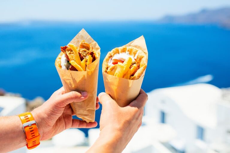 ヨーロッパの旅行先で手軽な食べ物として人気の、ギリシャ名物「ギロス」。青い海と白い建物が美しい景勝地を背景に、お肉やフライドポテトがたっぷり詰まったボリューミーなピタパンサンドを手に掲げている様子