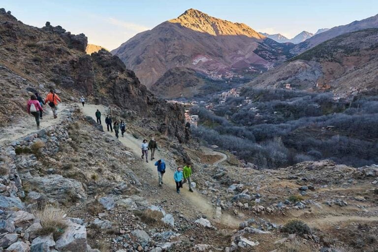 壮大な山々と眼下に広がる村の景色を楽しみながら、モロッコ・アトラス山脈の岩肌が続く登山道をハイキングする人々の様子