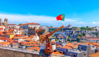 ヨーロッパで安く海外旅行できる国の代表格であるポルトガルにて、オレンジ屋根が連なるポルトの美しい街並みを背景に国旗を掲げて景色を楽しむ女性旅行者の後ろ姿