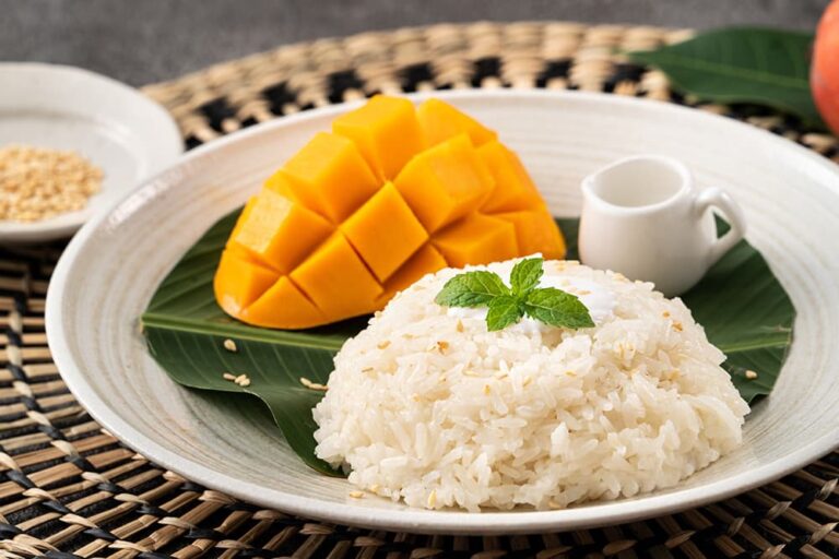 白い器のバナナの葉の上に美しく盛り付けられた、タイの定番の食べ物であるスイーツ「マンゴースティッキーライス」。大きくカットされた新鮮なマンゴーと、ミントを添えたもち米の様子