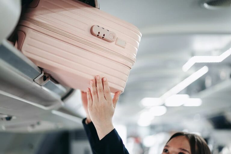 飛行機の機内で、頭上の収納棚に機内持ち込みサイズのピンク色のスーツケースを収納する女性旅行者