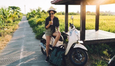 バリ島の田園地帯で、スクーターにまたがりスマートフォンで移動ルートや配車アプリを確認する旅行者の女性