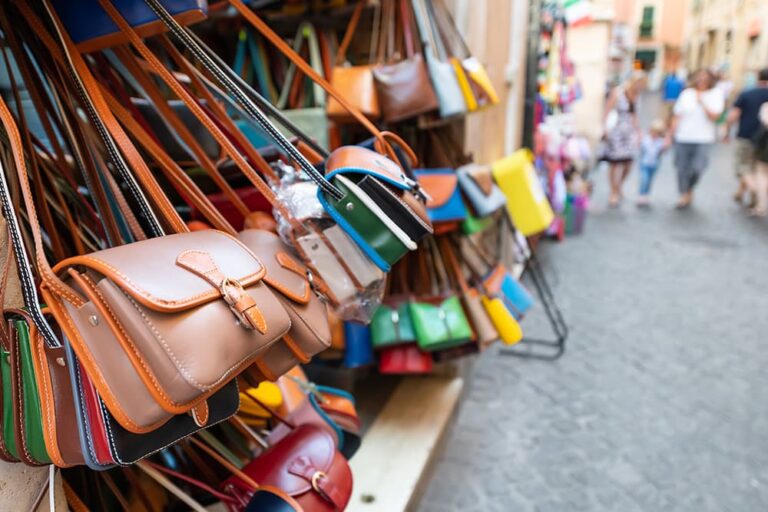 ローマのお土産として人気のイタリアンレザー製品。石畳の路地に面したショップの店頭に、色鮮やかな革のショルダーバッグが多数吊るされて販売されている様子