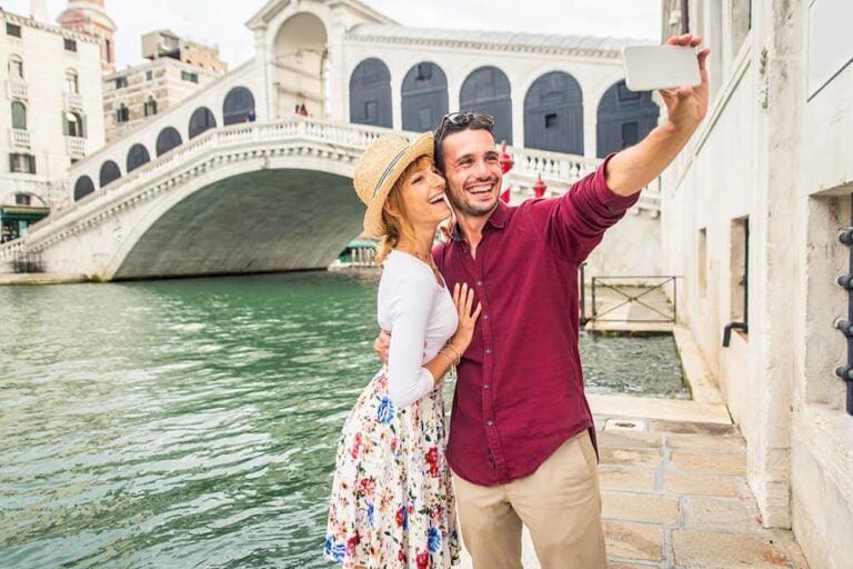 イタリア・ヴェネツィアの歴史的な橋と運河を背景に、新婚旅行で街歩きを楽しみながら笑顔で自撮りをするカップル