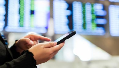 記事「Visit Japan Webの登録方法と帰国時の使い方」のサムネイル画像。空港のフライト案内板を背景にスマートフォンを操作する様子