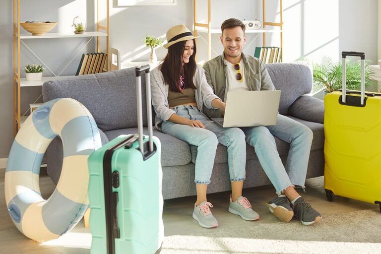 スーツケースなどの旅行アイテムを傍らに置き、ノートパソコンを見ながら2人に合う新婚旅行の行き先や日程を笑顔で相談して決めている様子
