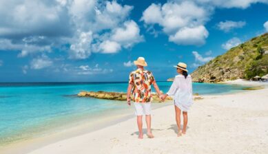 青く澄み渡る海と白い砂浜が広がる海外リゾートで、手をつないでビーチを歩く新婚カップルの後ろ姿