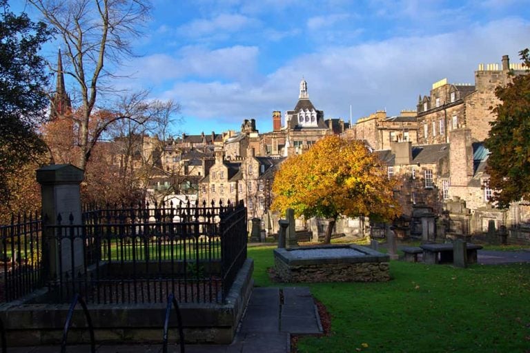 歴史的な石造りの街並みと秋の紅葉を背景にした、エディンバラのグレーフライアーズ・カークヤードの風景。「誓約派の牢獄」の歴史を伝える、鉄柵で囲まれた墓所が見えます。