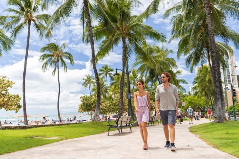 ハワイ・オアフ島にて、ヤシの木が並ぶビーチ沿いの道を笑顔で散策する新婚カップルの様子