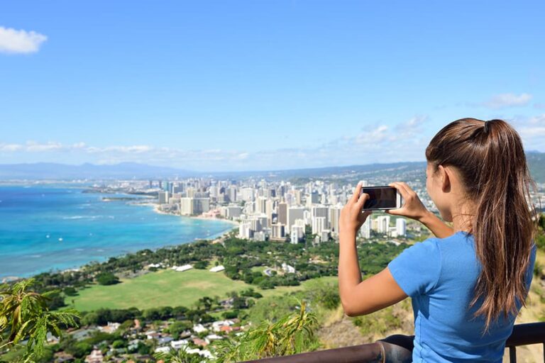青い海とワイキキの街並みを一望できるオアフ島の高台から、スマートフォンで風景を撮影する女性の後ろ姿