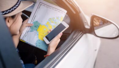 車内で紙の地図とスマートフォンを手に持ち、海外旅行先でルート検索やデータ通信を行う女性の様子