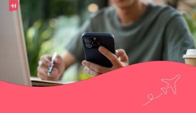 لقطة مقربة لرجل ينظر إلى هاتفه الذكي في يده بينما يد أخرى تمسك بقلم وتكتب في دفتر ملاحظات وهو جالس على طاولة خشبية.