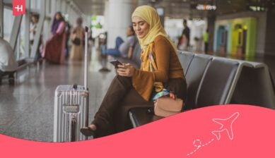 امرأة مسافرة تجلس في المطار وتستخدم هاتفها الذكي وبجانبها حقيبة سفر، مما يعبر عن أهمية البقاء على اتصال عبر التجوال الدولي أو شريحة eSIM أثناء التنقل.