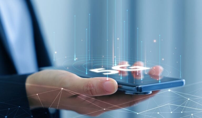 يد تحمل هاتفاً ذكياً يظهر عليه أيقونة شبكة الجيل الخامس (5G) وواجهة شبكة عالمية.