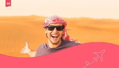 سائح شاب سعيد ومتحمس يقف باسطاً ذراعيه، وخلفيته كثبان رمال الصحراء العربية الذهبية.