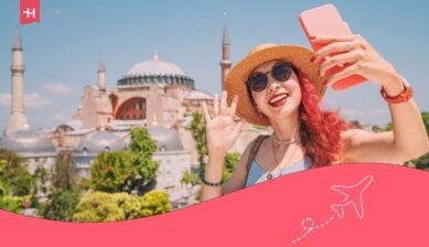 En smilende kvinne med hatt og solbriller tar en selfie foran Hagia Sofia i Istanbul, Tyrkia. Under er et rosa grafisk element med et flyikon.
