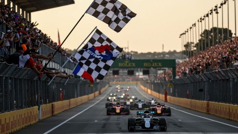 Formel 1-biler som raser nedover banen under en solnedgang med rutete flagg i forgrunnen.